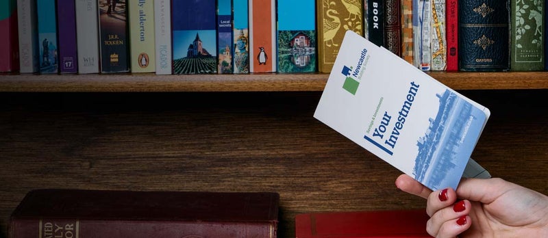 A close up of someone holding a Newcastle Building Society Passbook with a book shelve in the background. 