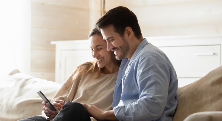 Happy man and woman sat on a sofa looking at a mobile phone, on a video call. 