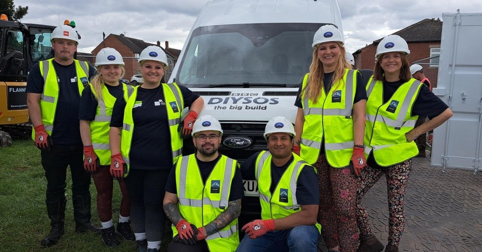 A group of Newcastle Building Society colleagues on a construction site, stood in front of a DIY SOS van, wearing high vis jackets and hard hats.