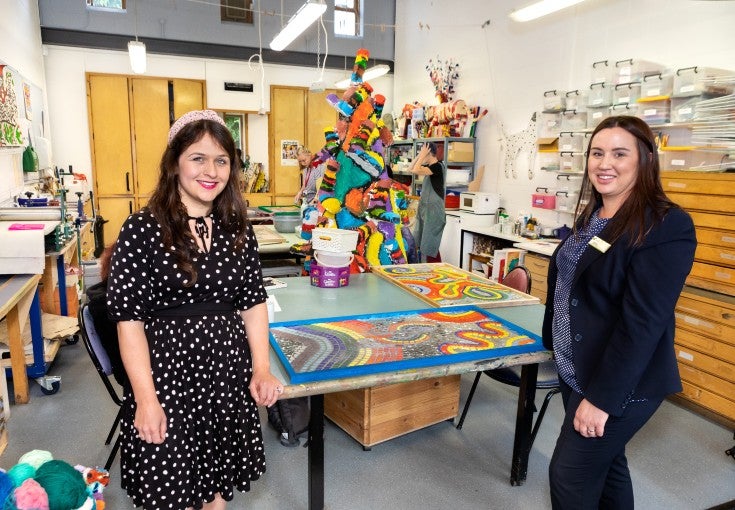 Two ladies stood beside a table, smiling for the camera. On top of the table is some mosaic art and in the background there is a colourful sculpture and two artists working. 