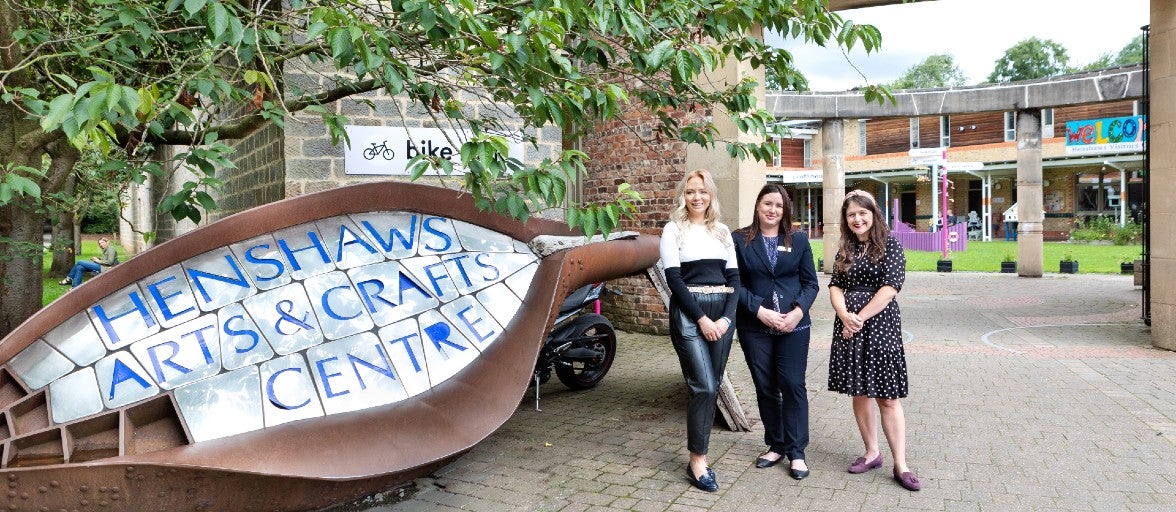 Three ladies stood outside of Henshaws & Crafts Centre. In the foreground on the left-hand side, is the large sign for the Centre and the ladies are stood to the right. The Centre is visible in the background.