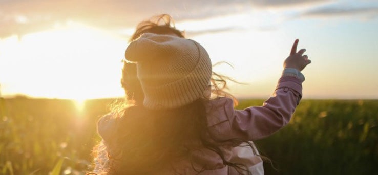 Mum holding her daughter, with their backs to us. The little girl is pointing to the sky. In the background there is a field and a sunset. 