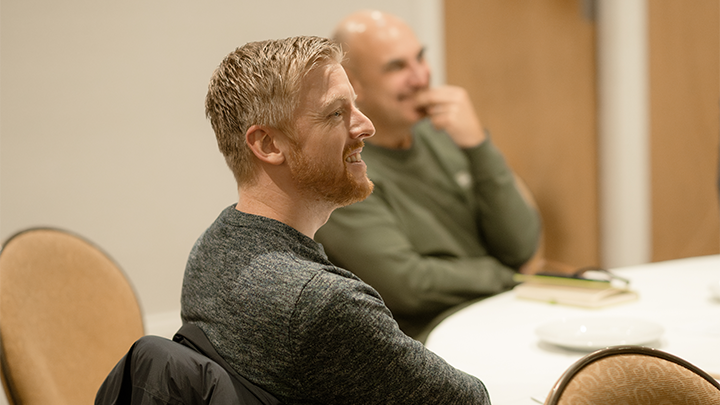 A man sitting at a table with another man in the background