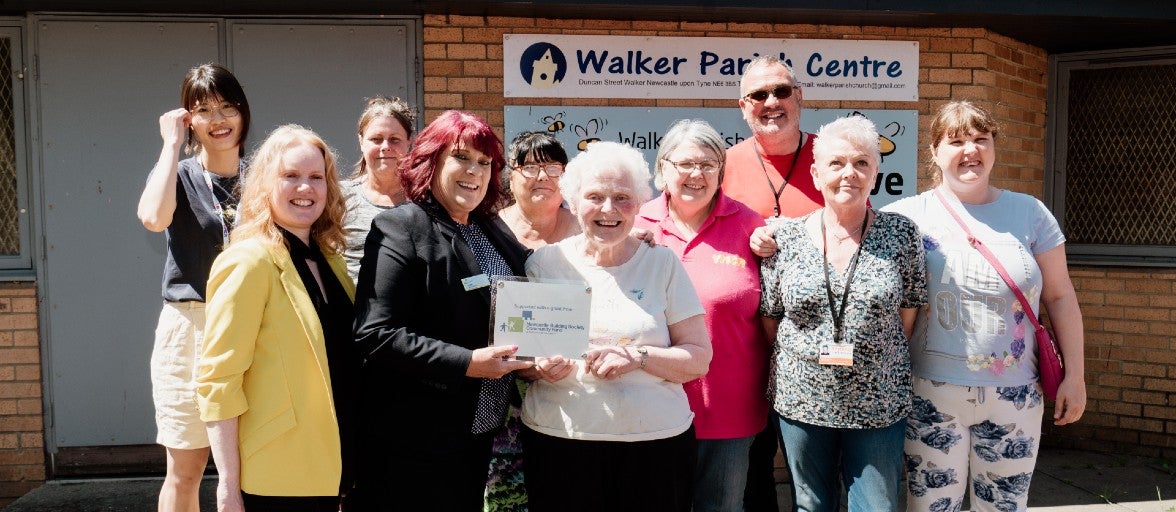 An over 55s friendship group stood outside a church, smiling for the camera, with two ladies at the front of the group holding a commemorative plaque. 