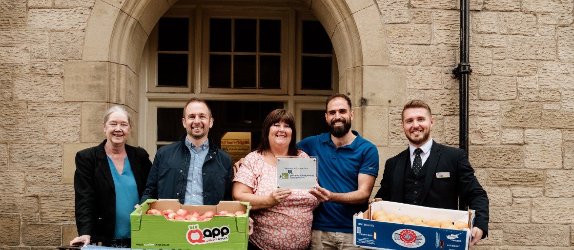 A group of people stood outside a building. Some of them are holding boxes of fruit and two of them are holding up a commemorative plaque together.