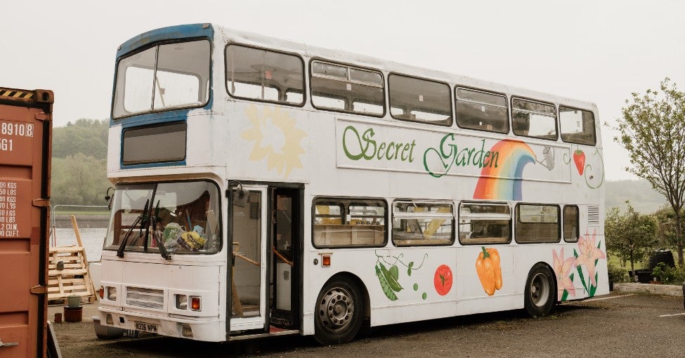 A white converted double decker bus. On the side of the bus reads "Secret Garden" and there is a rainbow and some fruit and vegetables painted on.