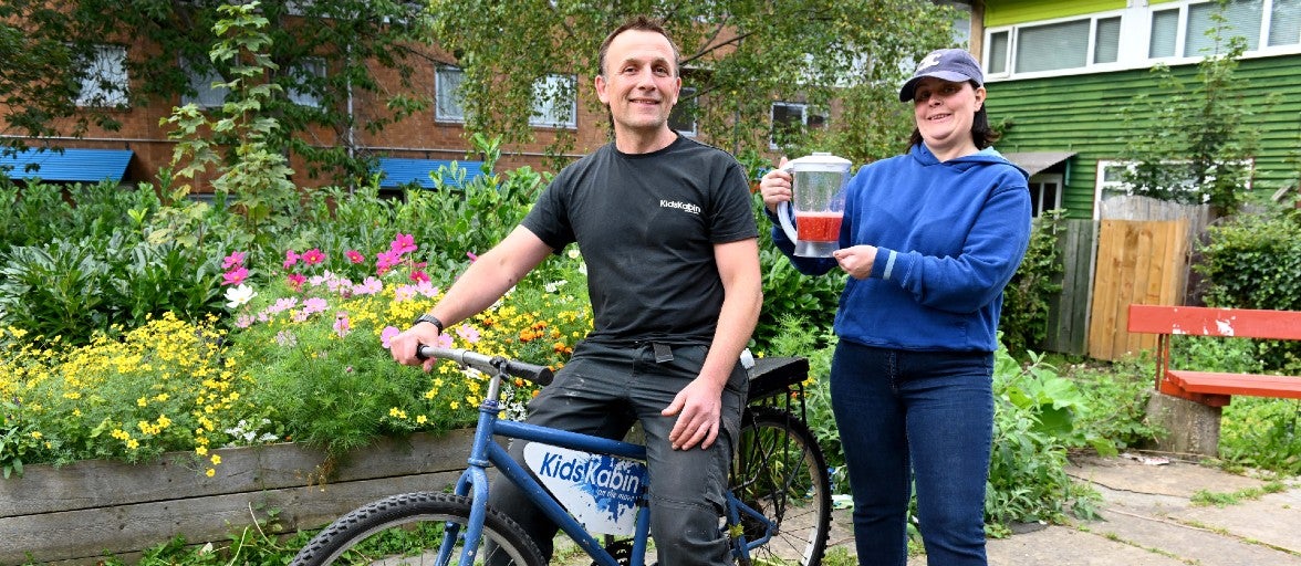 Will Benson, Chief Officer at Kids Kabin, sat on a bike to demonstrate how a bike-powered smoothie maker works. Behind him is Joanne Oskarsen-Stewart, our Head of Environment and Sustainability, holding a blender containing a smoothie.