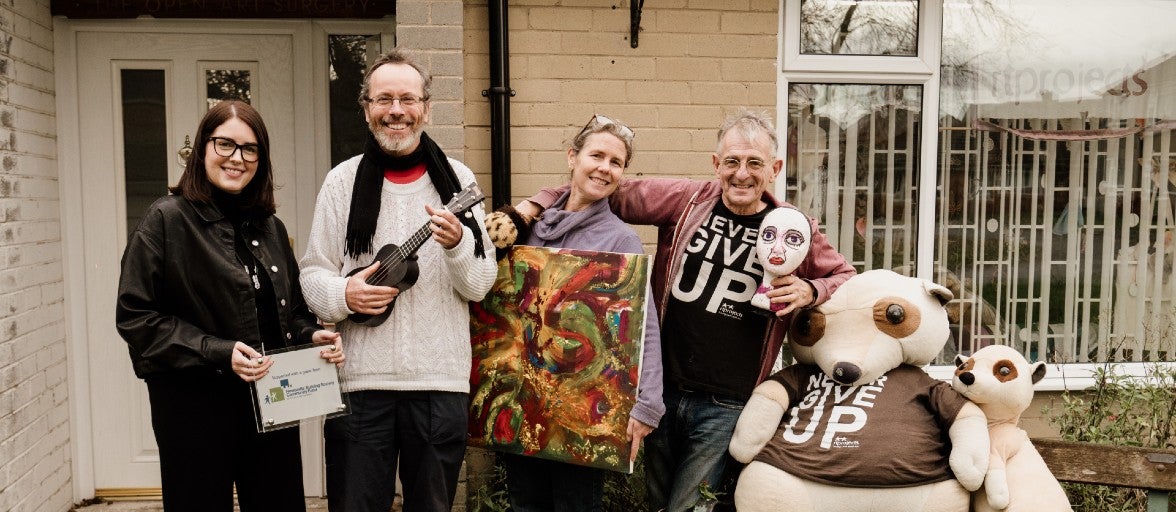 Two men and women stood outside of a house holding up pieces of art and instruments. Beside them are also two large plush bears.