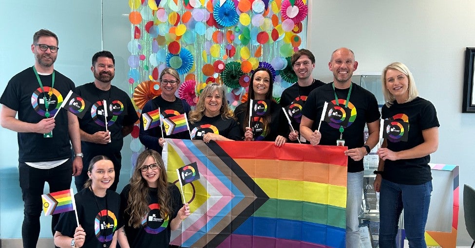 A group of colleagues wearing Pride t-shirts, each holding mini Pride flags and one large Pride flag.