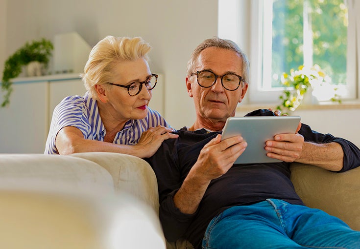 Elderly man sitting on sofa in the living room at home and showing something on digital tablet to his wife.