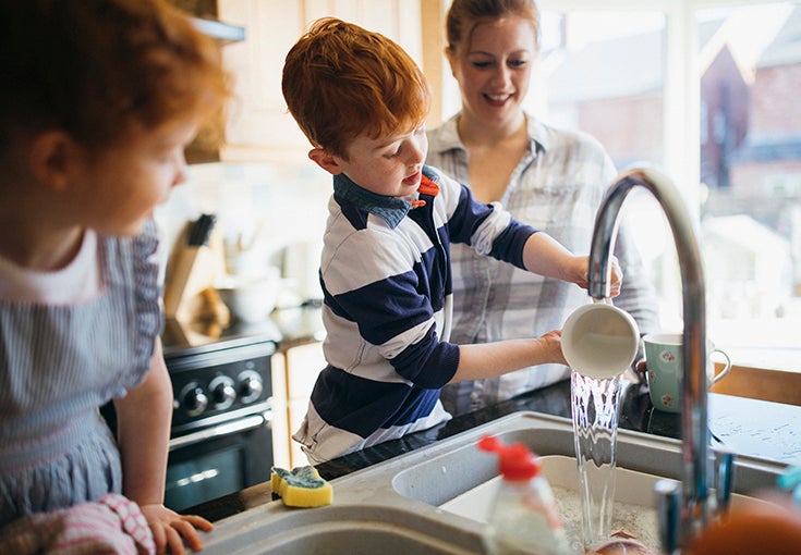 Two children and their mother washing the dishes in the kitchen sink.
