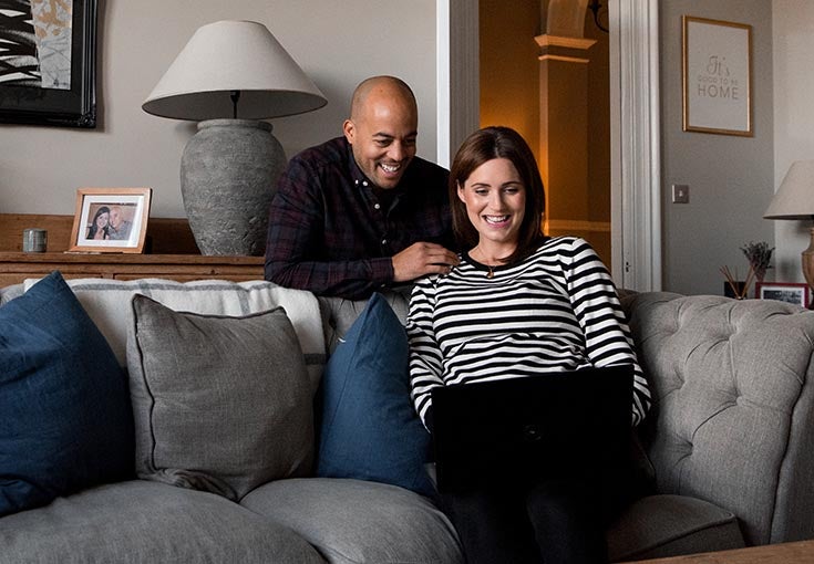 Man and woman sat in living room looking at a laptop smiling.