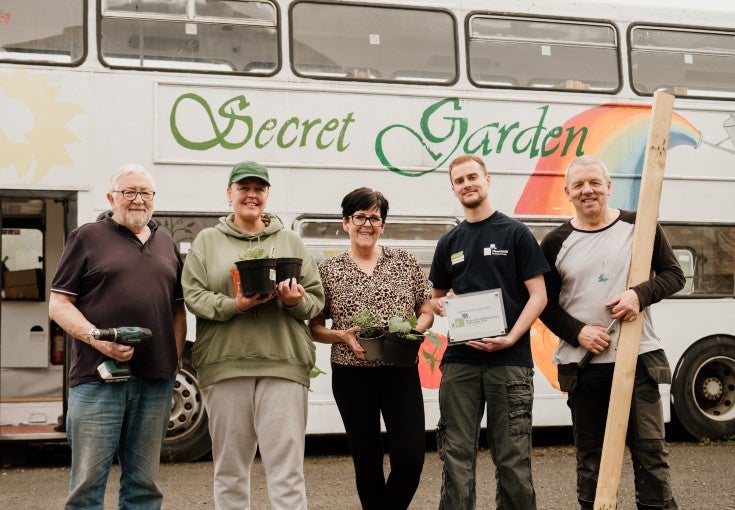 A group of people holding gardening stood beside a converted, white double decker bus.