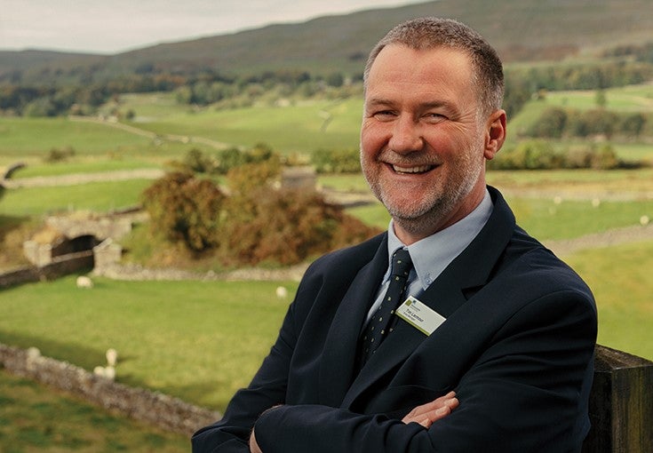 Man smiling with folded arms, with a scenic view of Hawes behind him. 