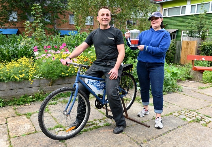 Will Benson, Chief Officer at Kids Kabin, sat on a bike to demonstrate how a bike-powered smoothie maker works. Behind him is Joanne Oskarsen-Stewart, our Head of Environment and Sustainability, holding a blender containing a smoothie. 