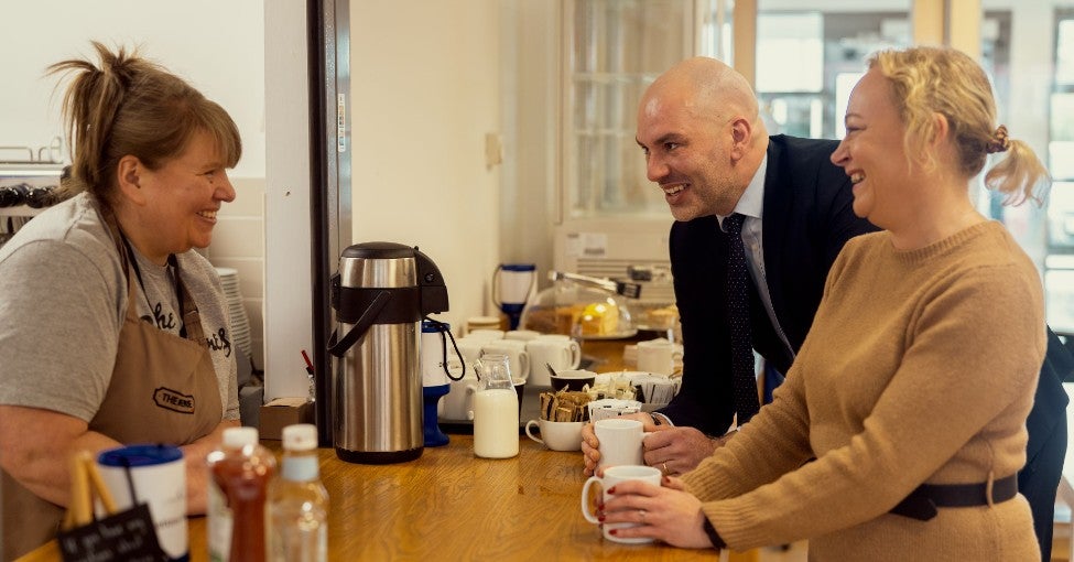 Three people stood together at a counter having a chat, laughing together, and enjoying cups of tea.