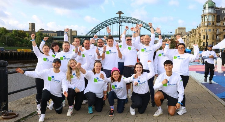 A group of Newcastle Building Society colleagues at Newcastle Quayside stood cheering beside the River Tyne, with the Tyne Bridge behind them. 