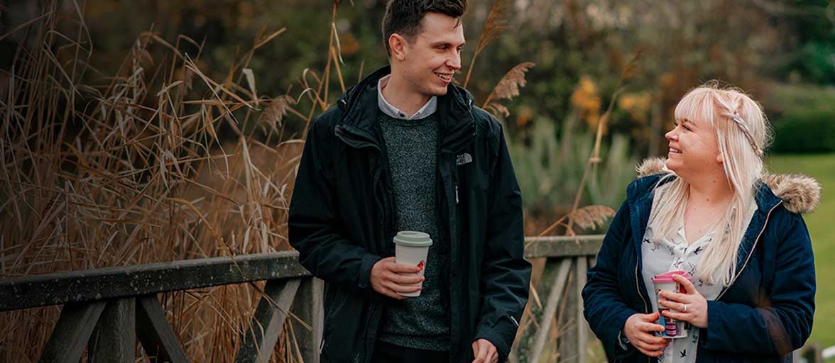 A man holding a cup and smiling while standing next to a fence, a woman looking at the man smiling and holding a cup. 