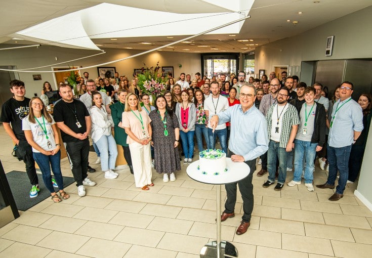 A group of around 80 office workers gathered together in the main reception of their office to celebrate their IIP Platinum reaccreditation. The man in their foreground is the Chief Executive Officer, Andrew Haigh, and he is cutting the celebration cake. 