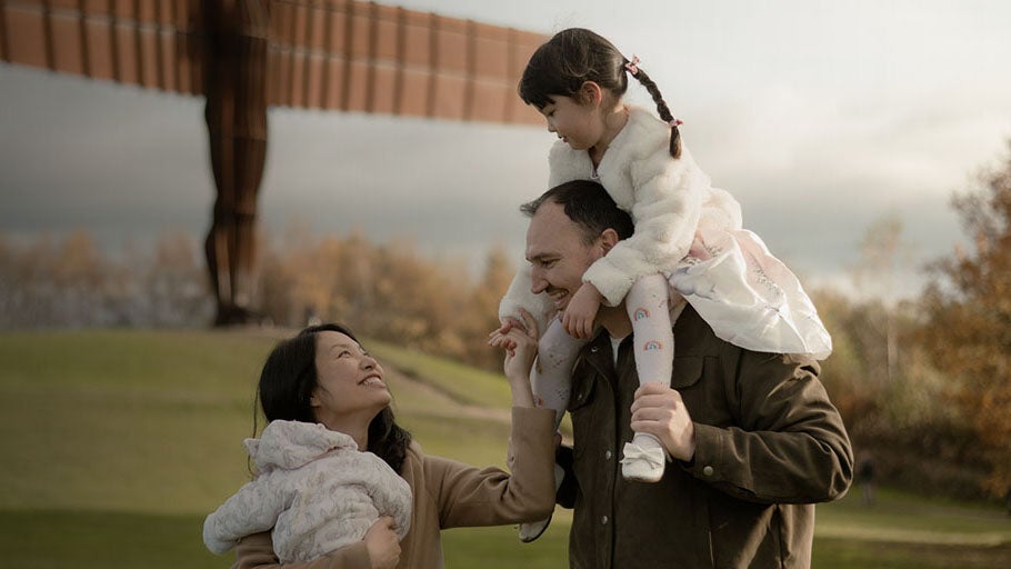 A family in front of the Angel of the North. A Mother, Father, a daughter on her Father's shoulders and a baby is held by her Mother. 