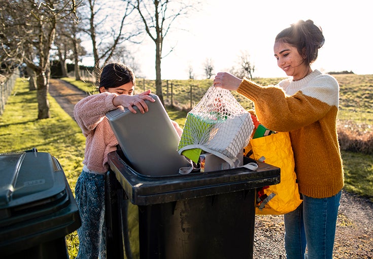 Two girls pouring their recycling waste into the bins out of bags at the bottom of their drive. 