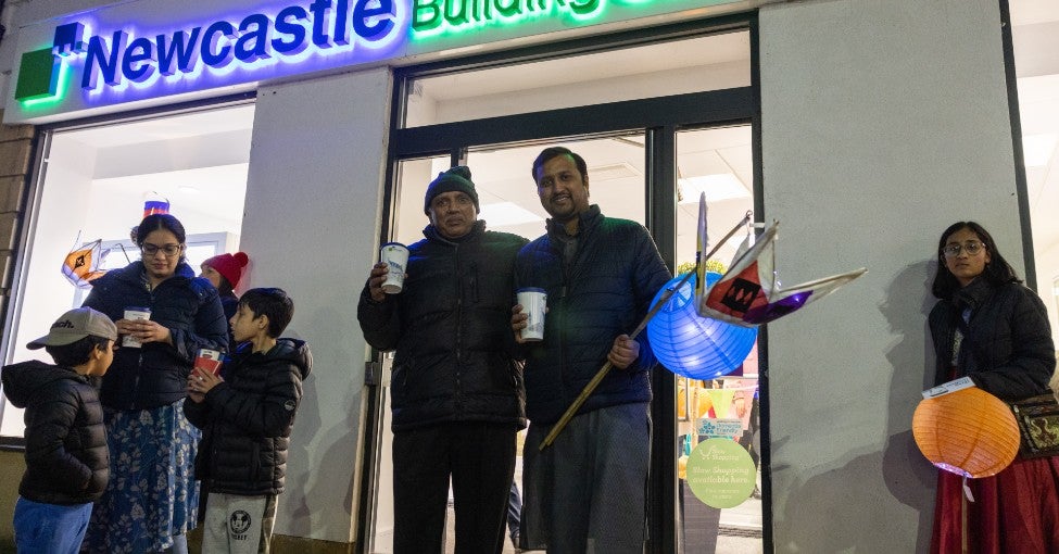 A group of people stood outside of a Newcastle Building Society branch at night, holding Diwali lanterns and reusable coffee cups. 