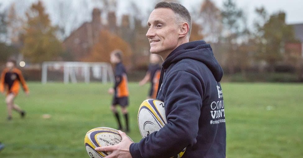 Close up of a man in a field holding two rugby balls in his hands.