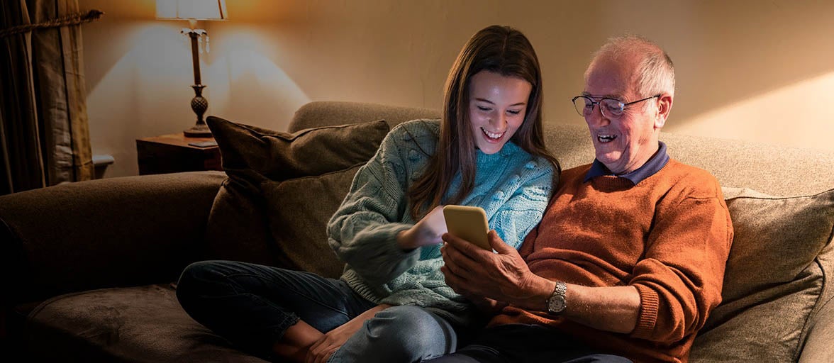 Senior man sat next to his teenage granddaughter on the sofa, looking at a mobile phone.