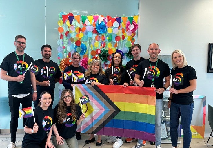 A group of colleagues wearing Pride t-shirts, each holding mini Pride flags and one large Pride flag.