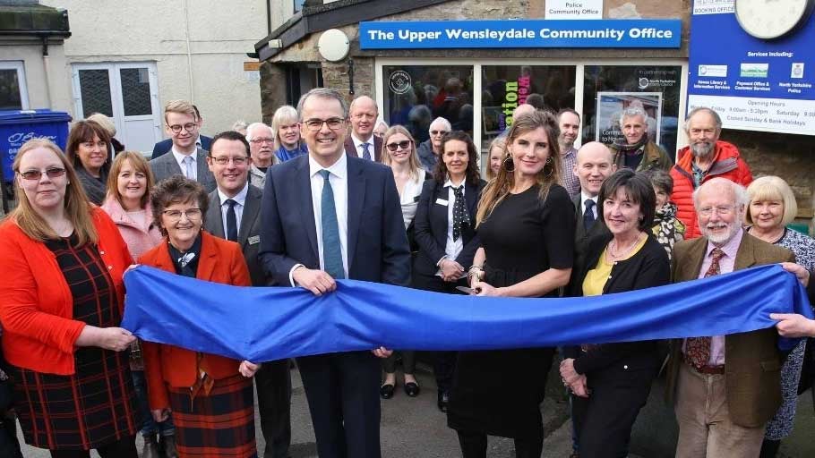 Exterior photo of the Hawes branch location within the Upper Wensleydale Community Centre. Various community members, customers and Newcastle Building Society colleagues all stood together holding a blue ribbon whilst a lady cuts it. 