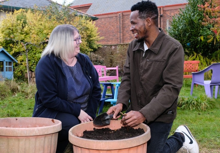 A man and woman facing each other and smiling, as they kneel beside a plant pot filled with soil.