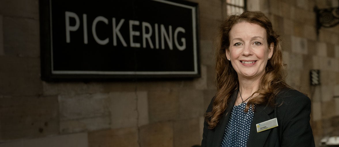 Pickering's Branch Manager, Vanessa, stood at the train station smiling with a sign reading 'Pickering', in the background. 