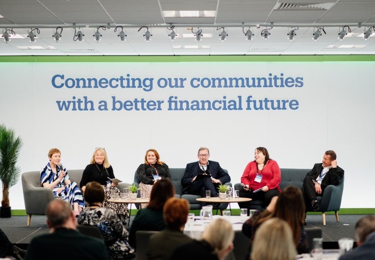 A panel of speakers on a stage, talking to a group of people sat at tables. In the background is Newcastle Building Society's purpose: "Connecting our communities with a better financial future". 