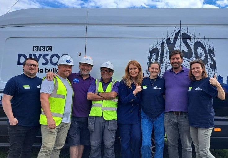A group of Newcastle Building Society colleagues stood with members of the DIY SOS team, including Nick Knowles, beside a DIY SOS van. 