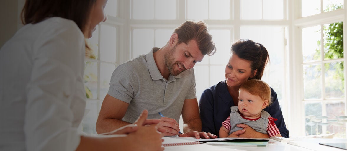 A Mum and Dad with their child sat at a table looking at documents, accompanied by a Financial Adviser. 