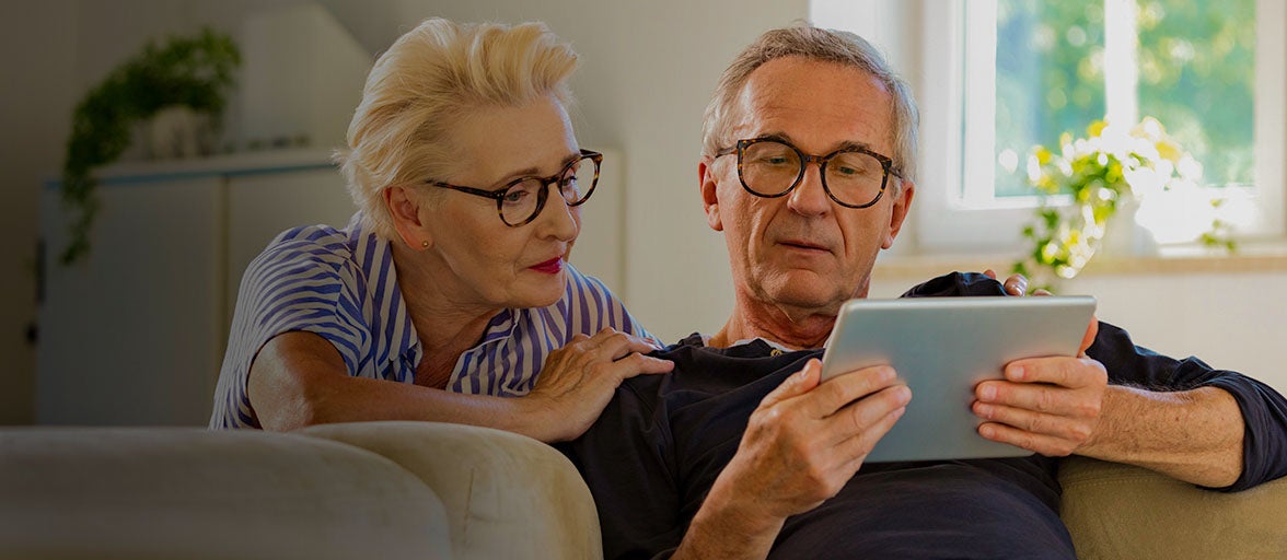 Man sat on sofa holding a tablet, with a woman kneeling down over his shoulder looking at the tablet. 