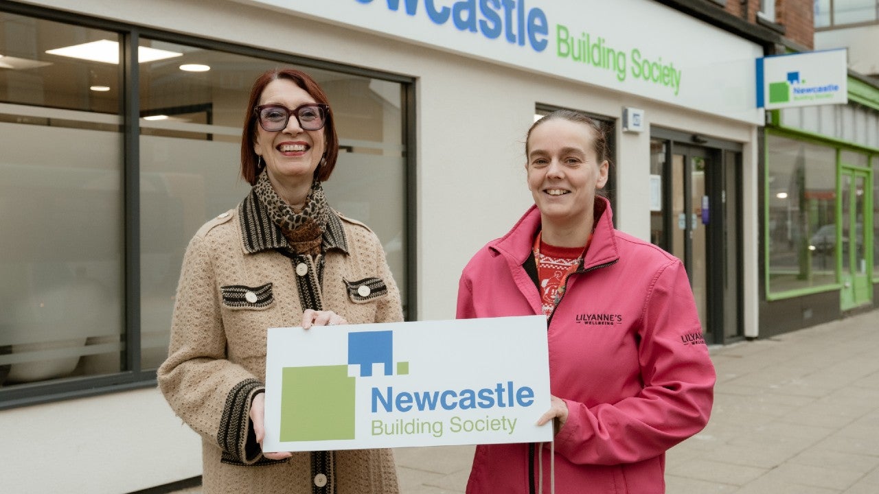 Two women stood outside of a Newcastle Building Society branch, holding a sign of the organisation's logo.