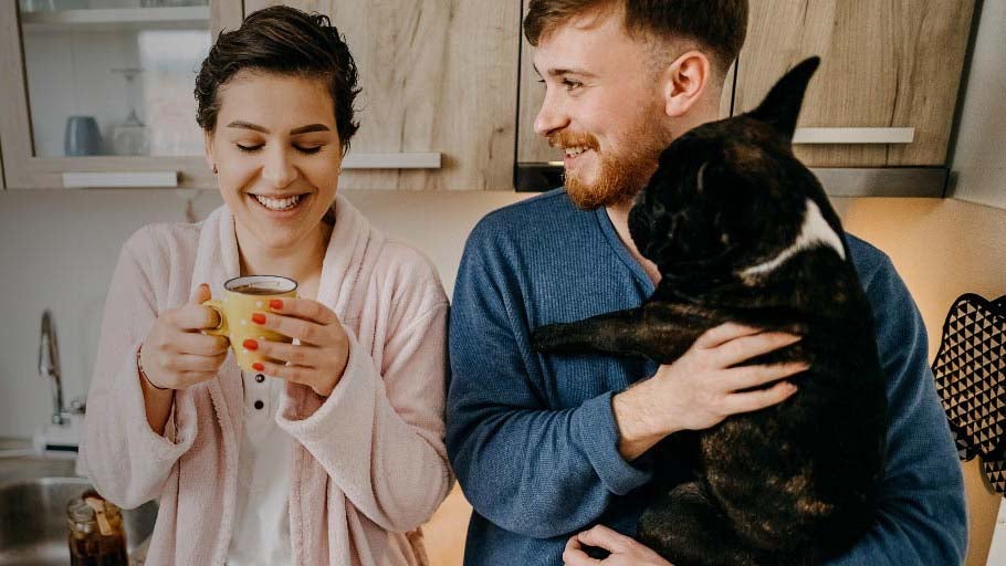 Woman and man in their kitchen having a cup of tea, man holding a dog. Both smiling. 