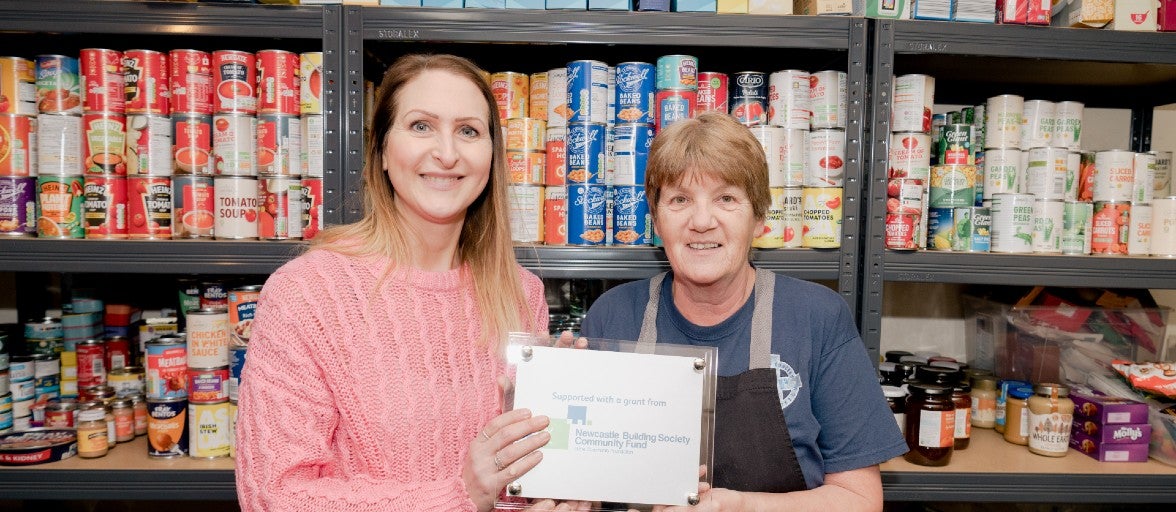 Suzanne Main, Head of Client Services at Newcastle Strategic Solutions - part of the Newcastle Building Society Group - stood in front of a shelf of food, holding a commemorative plaque with St Vincent's catering supervisor, Jan Cruikshanks.