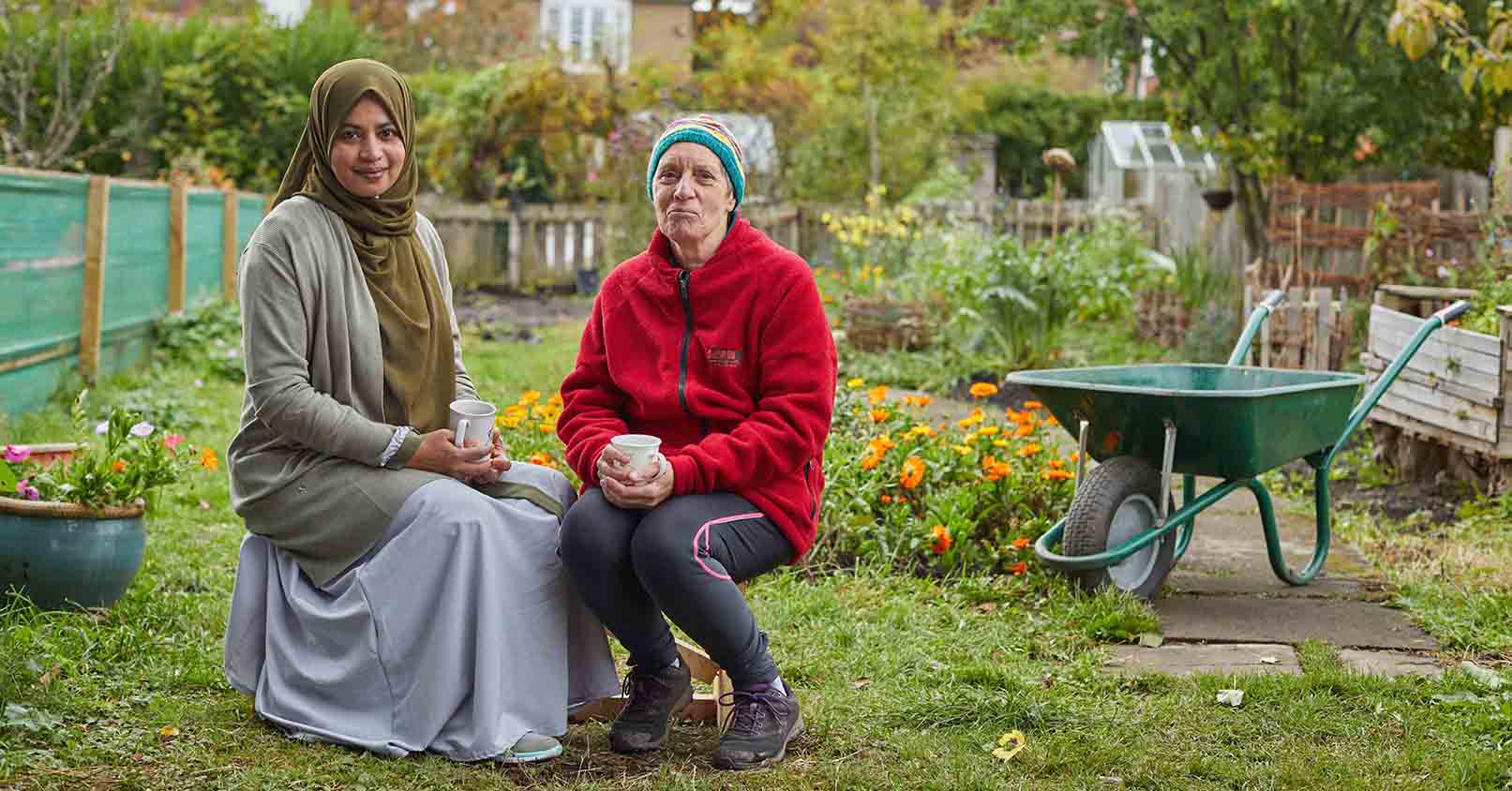 Two ladies sat in a community garden holding cups of tea. 