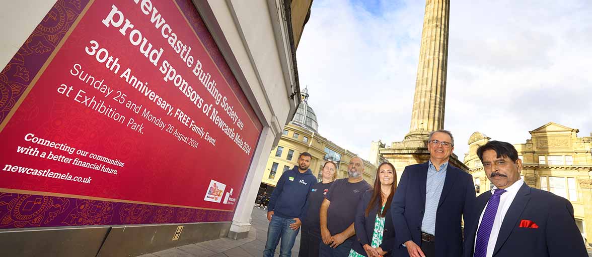 six people standing in front of Grey's Monument in Newcastle by the Newcastle Building Society branch window with a Newcastle Mela hoarding 