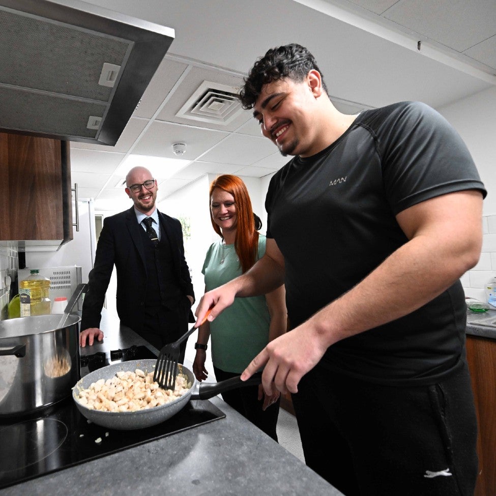 Three people stood beside a kitchen stove, smiling, as one man sautés chicken pieces in a frying pan.