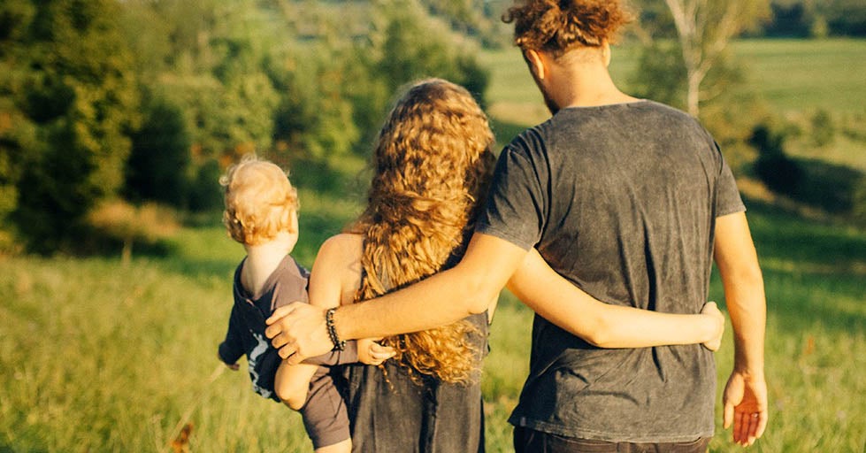 Family of three, a Mum, Dad and baby walking with their backs to us in a field. 