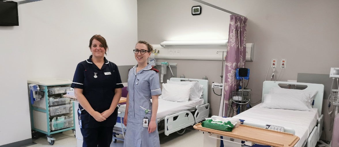 Two NHS staff members stood inside a hospital ward, with two empty hospital beds behind them. 