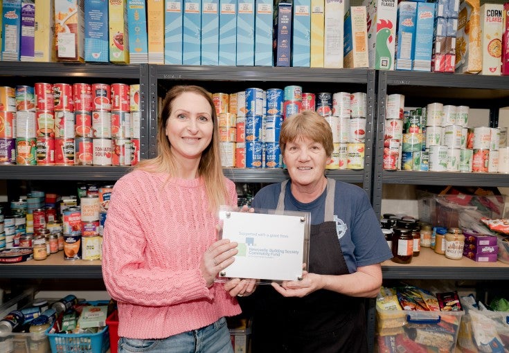 Suzanne Main, Head of Client Services at Newcastle Strategic Solutions - part of the Newcastle Building Society Group - stood in front of a shelf of food, holding a commemorative plaque with St Vincent's catering supervisor, Jan Cruikshanks.