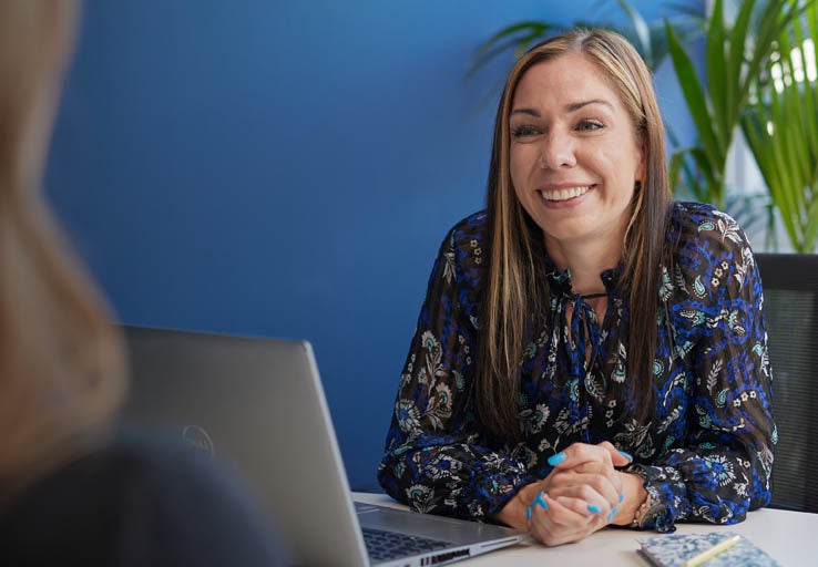 A woman smiling at another woman, as she sits behind a desk and a laptop on the desk. 