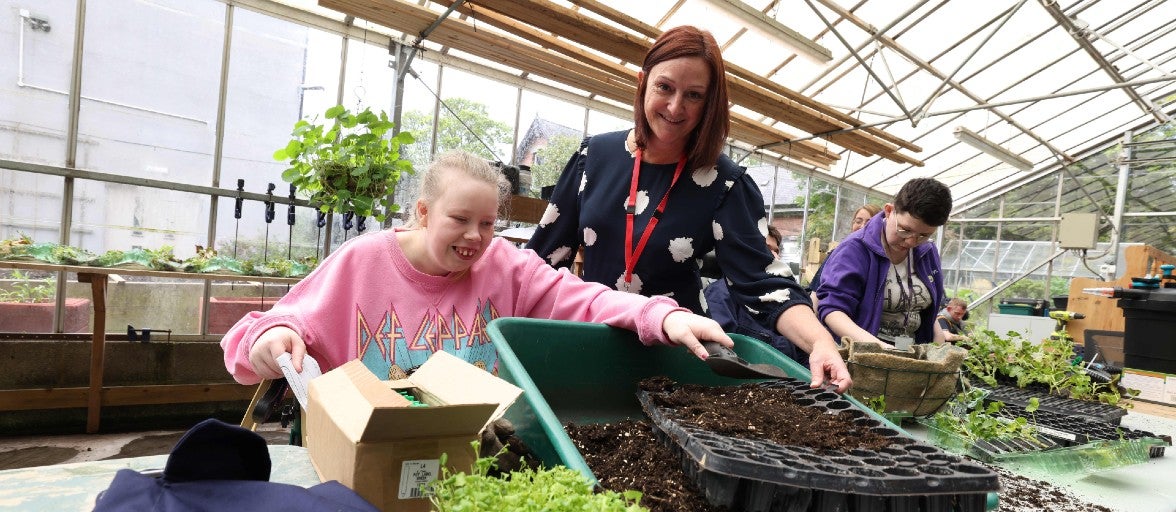 A lady and a child stood behind a table with some plant pots and plants. The lady is smiling at the camera and the young girl is reaching across to grab some soil and plant pots.