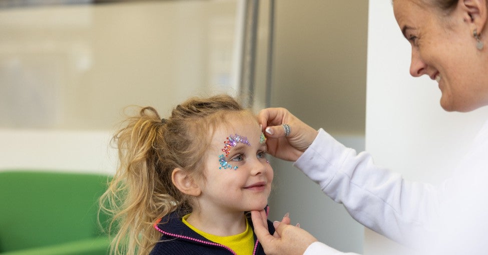 A little girl smiling up at a woman who is doing her face paint.