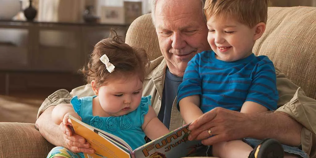 Granddad sitting on sofa reading childrens book to his grandkids as they sit on his lap