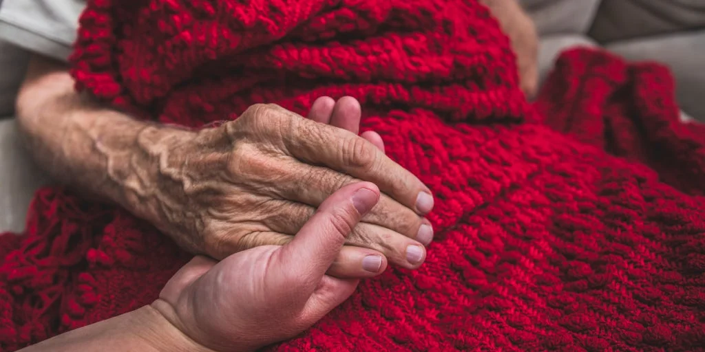 Close up child holding elderly parents hand as he sits under a red blanket